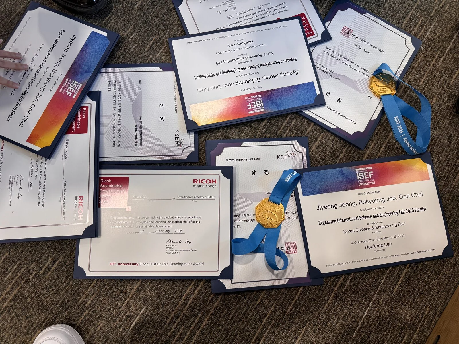 Award certificates and gold medals from Regeneron ISEF 2025 — naming Jiyeong Jeong, Bokyoung Joo and One Choi as finalists — alongside the Ricoh Sustainable Development Award and KSEF certificates.