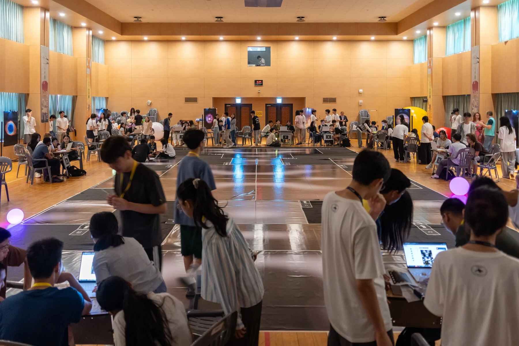 The competition hall, teams working at tables across a grid-marked floor under ambient lights.